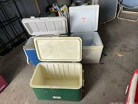 Three vintage coolers stacked and opened: green Thermos cooler, blue Coleman cooler, white Koolatron car-powered cooler. Shows general wear and use.