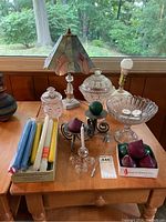 Table with small stained slag glass lamp with marble base, assorted colored taper candles, clear glass candle holders, covered glass compote, decorative glass jar with lid, a candle snuffer, and decorative red apples in a box.