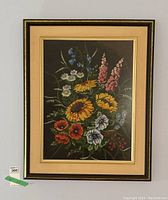Front view of framed needlepoint floral design showing vibrant multicolor flowers against dark background with beige matting and brown frame.