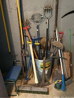 Photo shows various garden tools including rakes, broom, pickaxe, shovels, and a metal garbage can without a lid inside a cellar or basement space.
