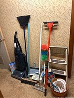 Miele upright vacuum cleaner and assorted cleaning tools including brooms, dustpan, mop, dusters, and step stool placed against patterned wall.