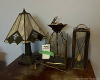 Photo showing three lamps on a wooden surface: a brass and slag glass desk lamp with trapezoidal shade on the left, a brass and glass hurricane lamp with etched glass panels and a brass candle snuffer in the middle, and a brass and glass lantern style lamp with a candle on the right.