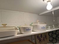 Shelf with three casserole dishes: two white CorningWare Cornflower Blue dishes with blue floral print and clear lids, plus a larger clear glass casserole dish with lid.
