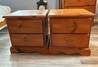 Front view of the pair of pine night stands showing wood grain, two drawers each with brass handles and scalloped base.