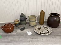 Group photo showing five pottery pieces including a bean pot with lid, small jar, tall bottle-shaped crock, large dark brown crock, and a flat decorative plate.