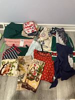 Overview of entire lot showing multiple folded table linens, aprons, oven mitts, and tea towels on a floor surface