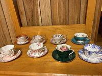 Photo showing the full set of eight vintage teacups and saucers on a wooden shelf spanning various brands and patterns.