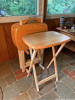 Four folding wooden TV tables stacked, with one unfolded showing light wood top and folding legs, near a window on tiled floor.