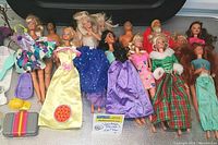 Wide view of all 13 Barbie and Ken dolls in various vintage dresses and outfits along with accessories on a silver reflective surface.