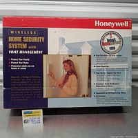 Full front facing box image showing branding, product name, summary of features, and a photo of a woman interacting with the security system device.