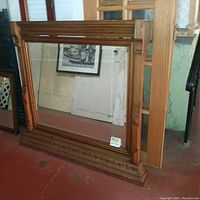 Full view of large oak wood mirror with beveled glass and mantel base sitting on floor.