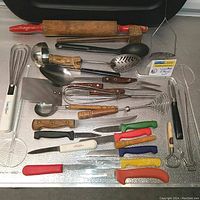 Photo showing assorted vintage kitchen utensils laid out on a silver metal surface including whisks, ladles, knives, peelers, and a wooden rolling pin with red handles.