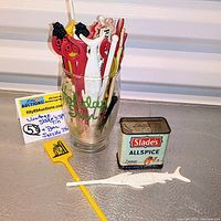 Photo showing the vintage Slade's Allspice tin placed next to a clear Holiday Inn glass filled with multiple colorful vintage bar picks and swizzle sticks, including a white fish-shaped swizzle stick lying flat in front.