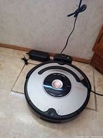 Photo of iRobot Roomba vacuum cleaner on floor connected to charging dock and power adapter, showing circular silver and black design with control button in center and visible use marks.