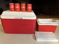 Large red Coleman cooler with white lid, small red Thermos cooler with separate lid, and four red Coleman drink holders placed on top of large cooler.
