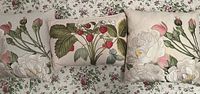 Three embroidered linen pillows arranged on a floral print surface showing detailed white flowers, pink buds, and strawberry embroidery.