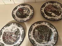 Four decorative plates showing various seasonal village scenes in brown and green tones with floral borders.