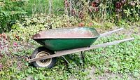 Side view of antique wheelbarrow showing green metal tray, wooden handles, and single rubber tire, with some rust and wear.