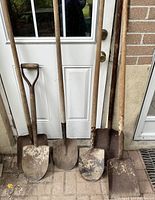 Photo showing six antique shovels with wooden handles and metal blades leaning against a white door and brick wall. Handles are worn and blades show rust and dirt.