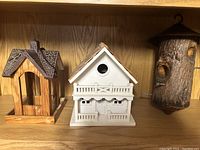 Photo of three wooden bird items on a wooden shelf: two bird feeders and one bird house.