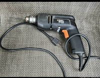 Side view of black Black and Decker electric drill with orange trigger and power cord connected, laid on fabric surface.