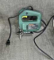 Bosch PST 50E electric jigsaw shown with green body, black power cord, yellow trigger, metal base, and blade attached, placed on a carpeted surface.