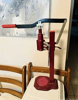 Red manual bottle capper with lever handle standing on a wooden chair against a wall.