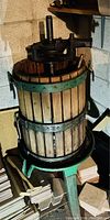 Front view of a large fruit wine press made of wood and cast iron, set against a concrete block basement wall with wooden boards nearby.
