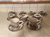 Top angle view of 7 teacups with matching saucers and 2 mugs with matching saucers arranged on a countertop, showing the Friendly Village pastoral pattern.