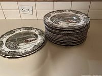 Stack of seventeen Friendly Village dinner plates showing winter village scene and decorative floral rim pattern.