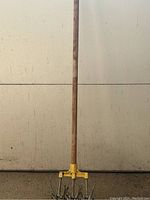 Full view of manual tiller showing long wooden handle and yellow metal head with rotating spiked tines for soil cultivation.