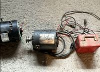 Two black electric motors with attached wires and pulleys, and a red rectangular device with handle and cables on concrete floor