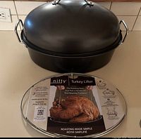 Photo showing large black turkey pot with domed lid and handles, with a circular metal turkey lifter roasting rack placed in front.