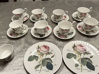 Set of six cups and saucers and three additional plates and bowl on a grey patterned tablecloth, showing floral rose design on all pieces.