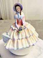 Front view showing young woman in bonnet and layered dress holding a bouquet, sitting on a bench which forms the trinket box lid.