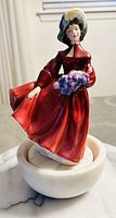 Front side view of the figurine showing facial expressions, the hat, and the lilac flowers she holds.