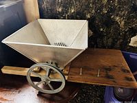 Full side view of the stainless steel manual grape crusher showing wide hopper, hand crank wheel, wooden handle, and mounting bolts.