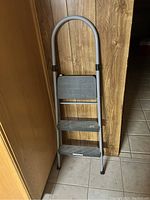 Front view of medium-sized 3-step metal ladder against wooden wall and tiled floor, showing rubber-tipped legs and black plastic treads.