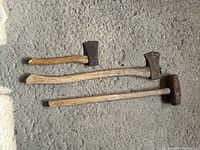 Photo of two axes and one sledgehammer laid on concrete floor showing worn wooden handles and rust on metal heads