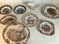 Top view of eight ceramic pieces featuring Friendly Village pattern including plates with various village scenes and an oval gravy boat.