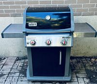 Closed lid view of Weber Spirit grill with side shelves extended, stainless steel control panel, red knobs and thermometer on lid.