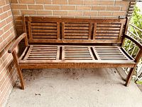 Full view of wooden outdoor bench showing slatted backrest and seat with armrests, wood has medium dark stain.