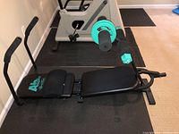 Side view of AbWorks abdominal exercise machine featuring padded bench, foam rollers, and resistance rings on black frame.