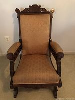 Front view of antique Victorian platform rocker with carved dark wood frame and beige patterned upholstery on seat, back and arms.