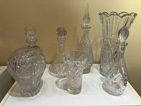 All seven crystal pieces consisting of three vases and four decanters arranged on a white surface with visible intricate cut patterns and designs.
