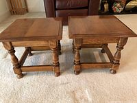 Pair of matching wooden end tables positioned on beige carpet with a brown leather chair in the background. Tables have a square top with beveled edges, turned legs, and stretcher supports connecting the legs.