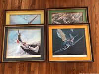 Four framed World War II fighter jet prints laid out on hardwood floor, showing detailed aircraft artwork and various colored mats and frames. One print has green background mat, another has yellow gold mat. All appear in wooden frames, one missing glass.