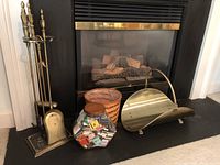 Photo showing brass fireplace tools and stand next to log caddy basket, glass jar filled with matchbooks, and coal scuttle.