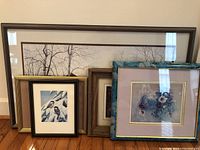 Five framed art pieces arranged leaning against a wall showing diversity in size, style, and subject matter including nature and floral themes.