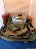 Photo showing stacked aluminum mess kit with lid and foldable cup on top of olive green duffel bag with surrounding items including seats and blankets.